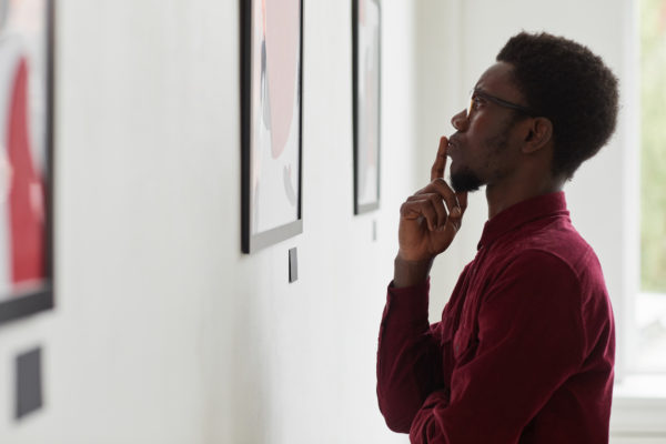 Side view portrait of young Black man looking at paintings and thinking at art gallery
